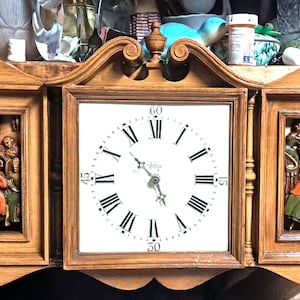 May include: A wooden wall clock with three sections. The center section features a white clock face with black Roman numeral hour markers and black hands. The left and right sections feature carved wooden scenes of musicians and dancers.