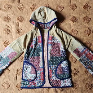 May include: A handmade, open-front jacket with a hood. The jacket features a patchwork design with various floral and geometric patterns in red, blue, green, and white. The sleeves and hood are a solid tan corduroy, and the jacket has two front pockets.