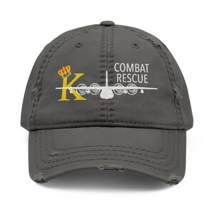May include: Distressed grey baseball cap with the text "COMBAT RESCUE" and a white airplane graphic. The cap also features a yellow "K" with a crown design.