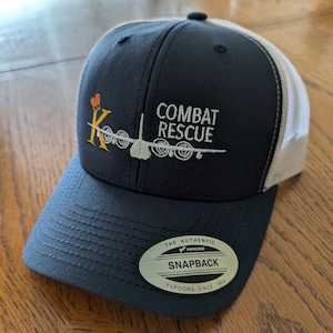 May include: A navy blue and white mesh baseball cap with an embroidered logo that says "Combat Rescue" and features a stylised airplane with a "K" in front of it. The cap has a white mesh back and a black "Yupoong" label that says "The Authentic Snapback Yupoong Since 1974".