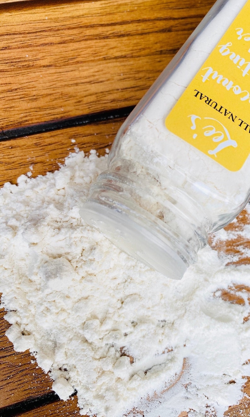 Puede incluir: Un frasco de vidrio transparente con una etiqueta amarilla que dice "Lee's ALL NATURAL Coconut" est&aacute; volcado, derramando polvo blanco sobre una superficie de madera. El polvo est&aacute; esparcido alrededor del frasco.