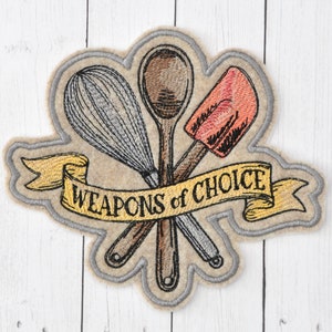 May include: Embroidered patch featuring a whisk, wooden spoon, and spatula, with the text "WEAPONS of CHOICE" on a banner. The patch has a light tan background with a gray border, perfect for a baker or chef.