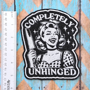 May include: Black and white embroidered patch featuring a vintage illustration of a woman holding a knife, with the words "COMPLETELY UNHINGED" in a bold font. The patch is approximately 4 inches wide.
