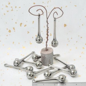 May include: Ten silver teardrop-shaped glass ornaments with a long, thin stem. The ornaments are displayed on a wooden stand and a ruler.