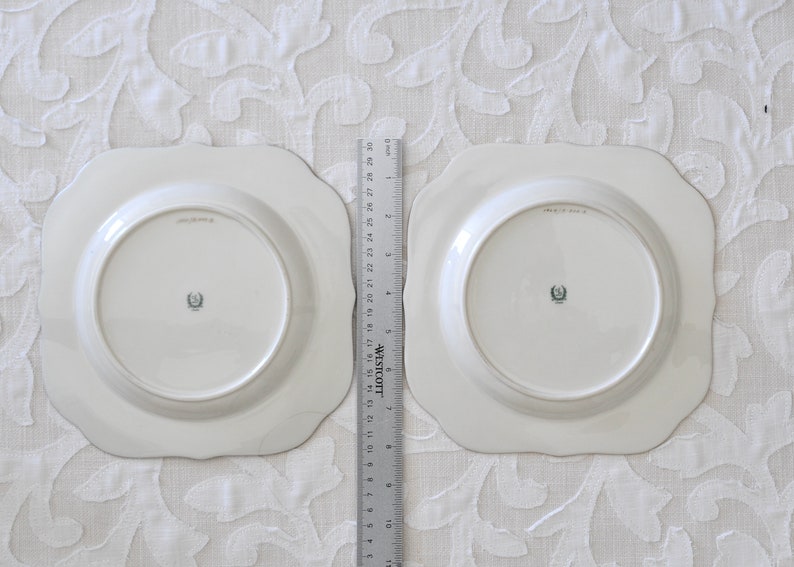 May include: Two white ceramic plates with scalloped edges. The plates have a raised rim and a center well. The back of each plate has a green and white logo.