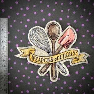 May include: An embroidered patch featuring a whisk, a wooden spoon, and a pink spatula arranged behind a yellow banner with the text WEAPONS of CHOICE. A metal ruler shows the patch is approximately 4.5 inches wide and 3.5 inches tall.