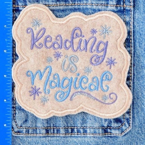 May include: A light beige embroidered patch with the words "Reading is Magical" in blue and purple script, accented with snowflake designs. The patch is cloud-shaped and sewn onto a denim pocket.