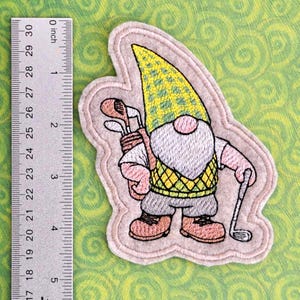 May include: Embroidered felt patch of a gnome wearing a yellow and green hat, a yellow and green vest, and holding golf clubs. The gnome is standing on a pink background.