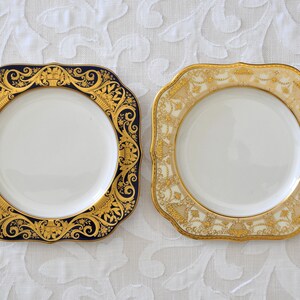 May include: Two square ceramic plates with gold trim. One plate has a blue background with a gold floral design. The other plate has a cream background with a gold floral design.