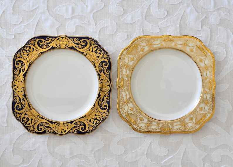May include: Two square ceramic plates with gold trim. One plate has a blue background with a gold floral design. The other plate has a cream background with a gold floral design.
