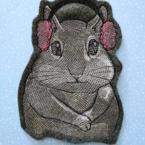May include: Embroidered gray felt patch of a gray hamster wearing pink earmuffs and a black headband.