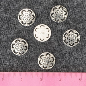 May include: Six silver metal buttons with intricate floral designs. The buttons are arranged in a circle on a gray fabric background. A ruler is visible at the bottom of the image.