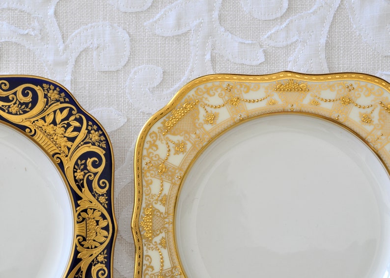 May include: Two decorative plates with gold and white designs. One plate has a blue rim and the other has a cream rim.
