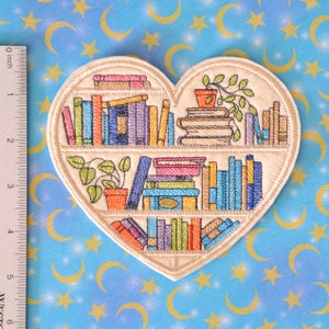 May include: Heart-shaped embroidered patch featuring a bookshelf design. The patch showcases a cream-colored heart with a bookshelf filled with colorful books, potted plants, and a cream-colored border. The patch measures approximately 4 inches in height.