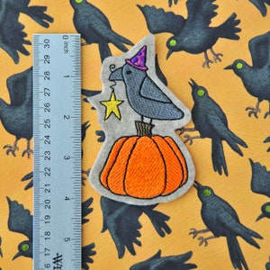 May include: A felt applique of a crow wearing a purple and pink witch hat, holding a gold star, perched on top of an orange pumpkin. The applique is on a yellow felt background with black crows.