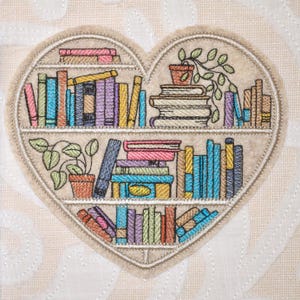 May include: Embroidered heart-shaped design featuring a bookshelf filled with colorful books and potted plants. The design includes books in various colors like pink, blue, and yellow. The background is a light beige fabric with a subtle pattern.