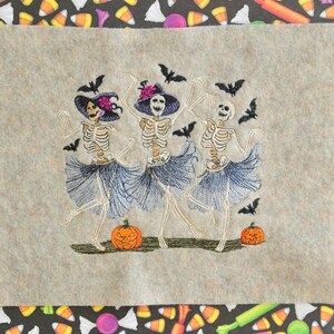 May include: Three skeleton figures wearing witch hats and tutus dance with bats flying around them.  Two orange pumpkins are on the ground.