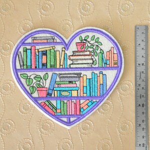 May include: A heart-shaped embroidered patch featuring three shelves of colorful books and potted plants. The design is outlined in purple thread on a white background. A metal ruler placed to the right shows the patch is approximately 5.5 inches tall.