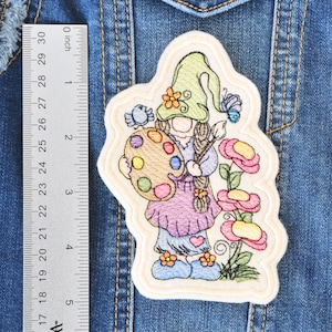 May include: Embroidered patch of a gnome holding a paint palette with colorful paint. The gnome is wearing a green hat and a purple dress. There are flowers and a butterfly in the background.