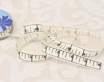 May include: A white fabric measuring tape with black markings. The tape is marked in inches from 1 to 6. The tape is rolled up and the end is visible.