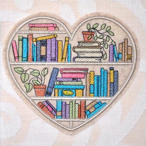 May include: A heart-shaped embroidered patch featuring three shelves filled with colorful books and potted plants. The design includes various shades of blue, yellow, pink, and green thread on a light beige fabric background with a stitched border.