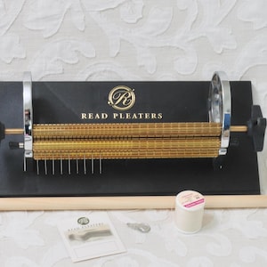 May include: A Read Pleaters machine featuring a black base with gold-colored rollers and silver side supports. The device includes a long wooden dowel, a small spool of white thread, a metal key, and a white instruction card with the brand logo.