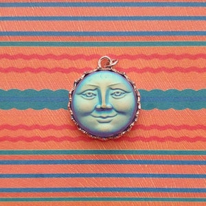 May include: A silver pendant with a blue and green iridescent face of the moon. The moon has a smiling expression with a round face and a small nose.