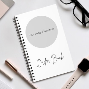May include: White spiral bound notebook with a gray circle in the center and the text "Your image / logo here". The text "Order Book" is written in cursive below the circle.