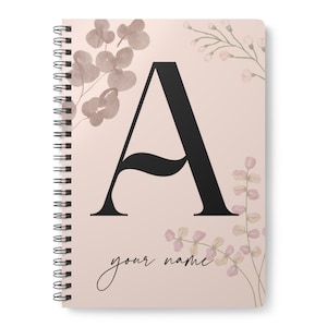 Puede incluir: Una libreta de espiral rosa con una letra A negra y diseños florales. El texto "your name" está impreso debajo de la letra A.