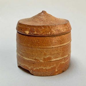 May include: A small, round, ceramic jar with a lid. The jar is a light brown color with a slightly rough texture. The lid has a pointed top.
