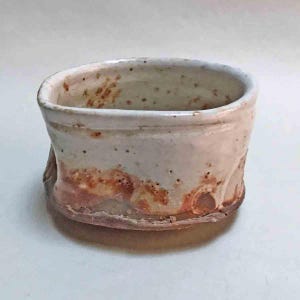 May include: A handmade ceramic bowl with a wide, oval shape. The bowl features a speckled, off-white glaze with reddish-brown accents around the base and interior. The rustic design gives it a unique, artisanal feel.