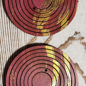 May include: Two round wooden coasters with concentric circles painted in shades of red and gold. The coasters have a hole in the center.