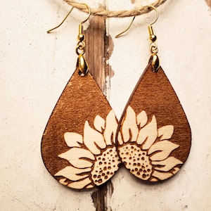 May include: A pair of teardrop-shaped wooden earrings with a laser-cut sunflower design. The earrings are brown with a light brown sunflower design. The earrings have gold hooks.