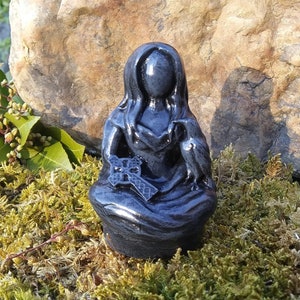 May include: A dark gray statue of a seated figure with long hair, holding a cross and a bird. The statue is set against a backdrop of moss and a large rock, with sunlight illuminating the details of the sculpture.