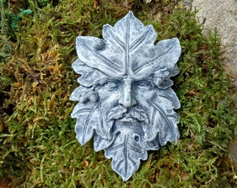 May include: A grey stone sculpture of a green man's face with leaves and acorns. The sculpture is set against a background of green moss.