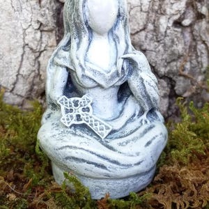 May include: A gray and white statue of a seated figure holding a cross and a bird. The figure has long hair and is wearing a draped garment. The statue is on a bed of green and brown moss.