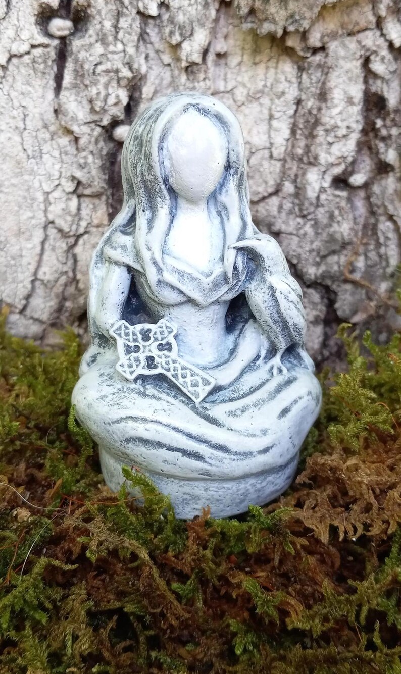 May include: A gray and white statue of a seated figure holding a cross and a bird. The figure has long hair and is wearing a draped garment. The statue is on a bed of green and brown moss.