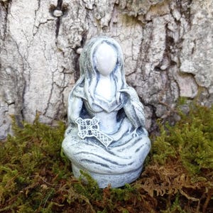 May include: A small, gray statue of a seated figure holding a cross and a bird. The figure is draped in flowing robes. The statue is set against a backdrop of tree bark and green moss.
