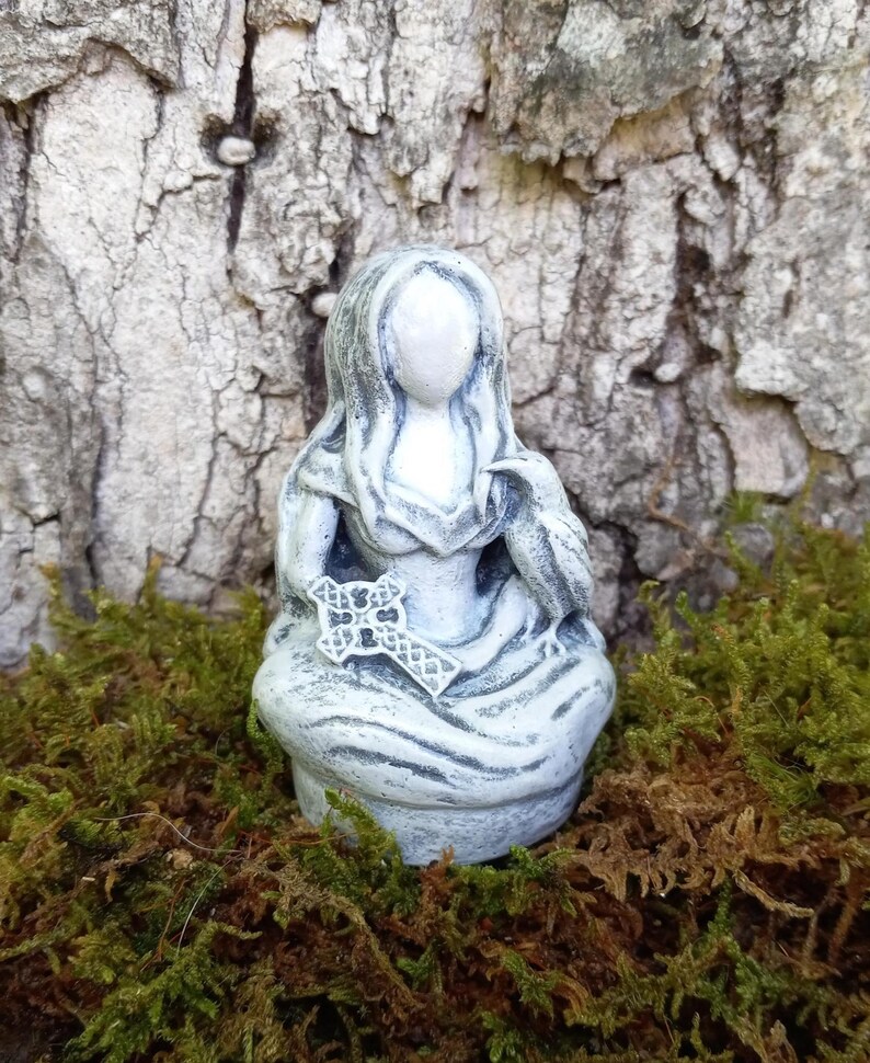 May include: A small, gray statue of a seated figure holding a cross and a bird. The figure is draped in flowing robes. The statue is set against a backdrop of tree bark and green moss.