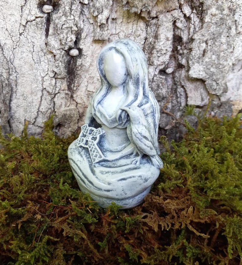 May include: A light blue statue of a seated figure with long hair, holding a bird and a cross. The statue is set against a backdrop of tree bark and green moss.