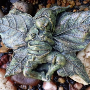 May include: A bronze-colored fairy figurine with leaf-shaped wings and a matching hat. The fairy is seated, with its hands clasped near its chest. The figurine has a textured finish and is surrounded by small rocks.