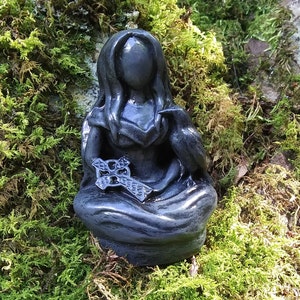 May include: A dark gray statue of a seated figure with long hair, holding a Celtic cross and a raven. The statue is set against a backdrop of green moss.