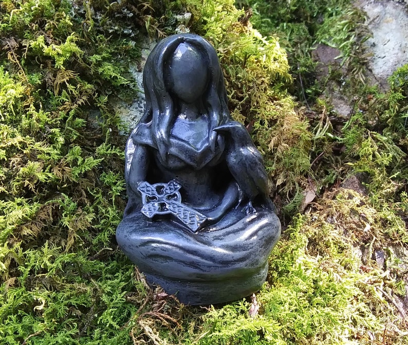 May include: A dark gray statue of a seated figure with long hair, holding a Celtic cross and a raven. The statue is set against a backdrop of green moss.