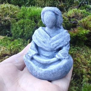 May include: A gray stone sculpture of a person sitting in a meditative pose. The sculpture is detailed with folds in the clothing and has a smooth, weathered finish.