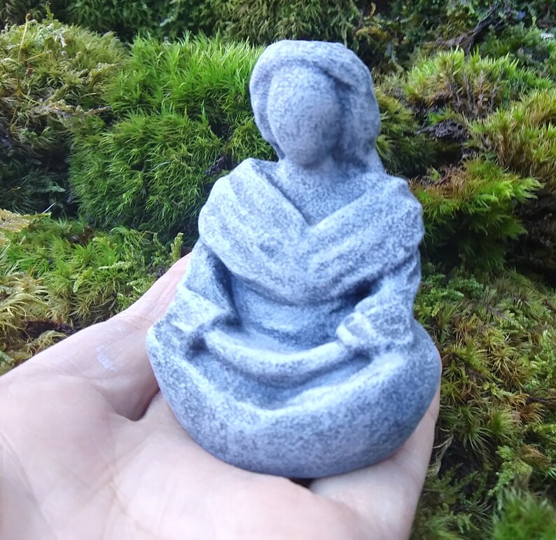 May include: A gray stone sculpture of a person sitting in a meditative pose. The sculpture is detailed with folds in the clothing and has a smooth, weathered finish.