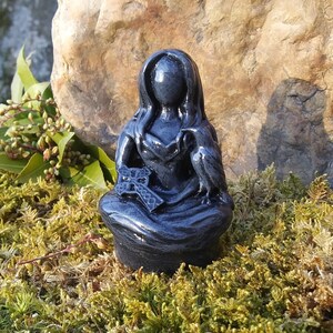 May include: A dark gray figurine of a seated figure with long hair and a hooded cloak. The figure holds a cross and has a bird perched on its shoulder. The statue is set against a natural background of moss and a large rock.