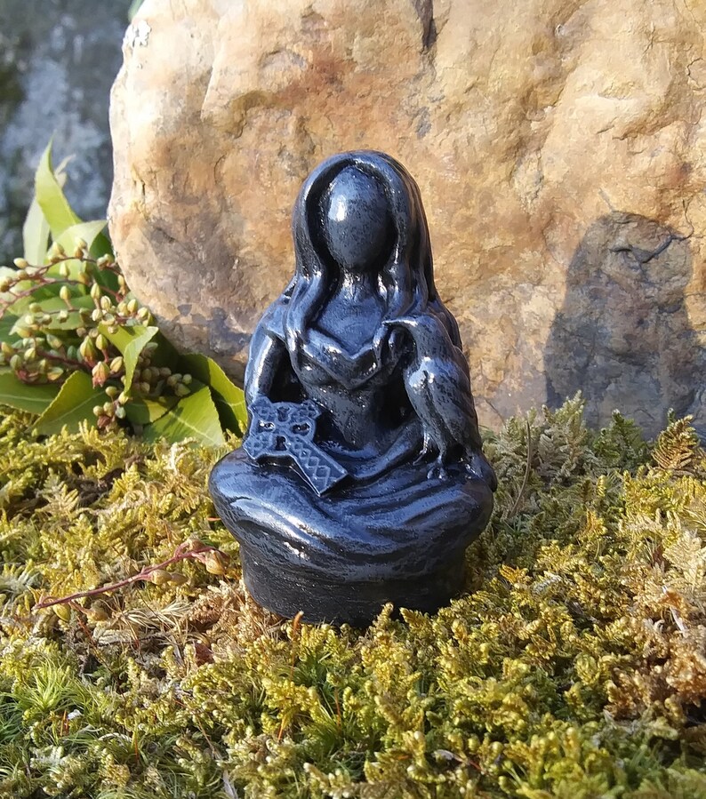 May include: A dark gray figurine of a seated figure with long hair and a hooded cloak. The figure holds a cross and has a bird perched on its shoulder. The statue is set against a natural background of moss and a large rock.