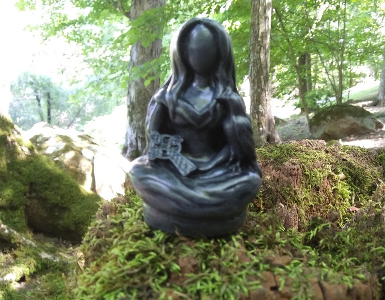 May include: A dark gray statue of a seated figure with long hair, holding a bird and a key. The statue is set on a moss-covered tree stump in a forest setting. The figure is wearing a long dress.