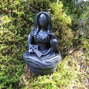 May include: A dark gray statue of a seated figure with long hair, holding a cross and a bird. The statue is set against a backdrop of green moss, creating a natural and mystical aesthetic. The figure's face is smooth, and the details of the cross are visible.