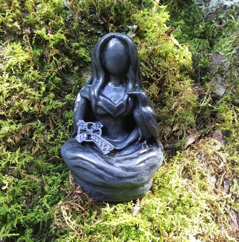 May include: A dark gray statue of a seated figure with long hair, holding a cross and a bird. The statue is set against a backdrop of green moss, creating a natural and mystical aesthetic. The figure's face is smooth, and the details of the cross are visible.